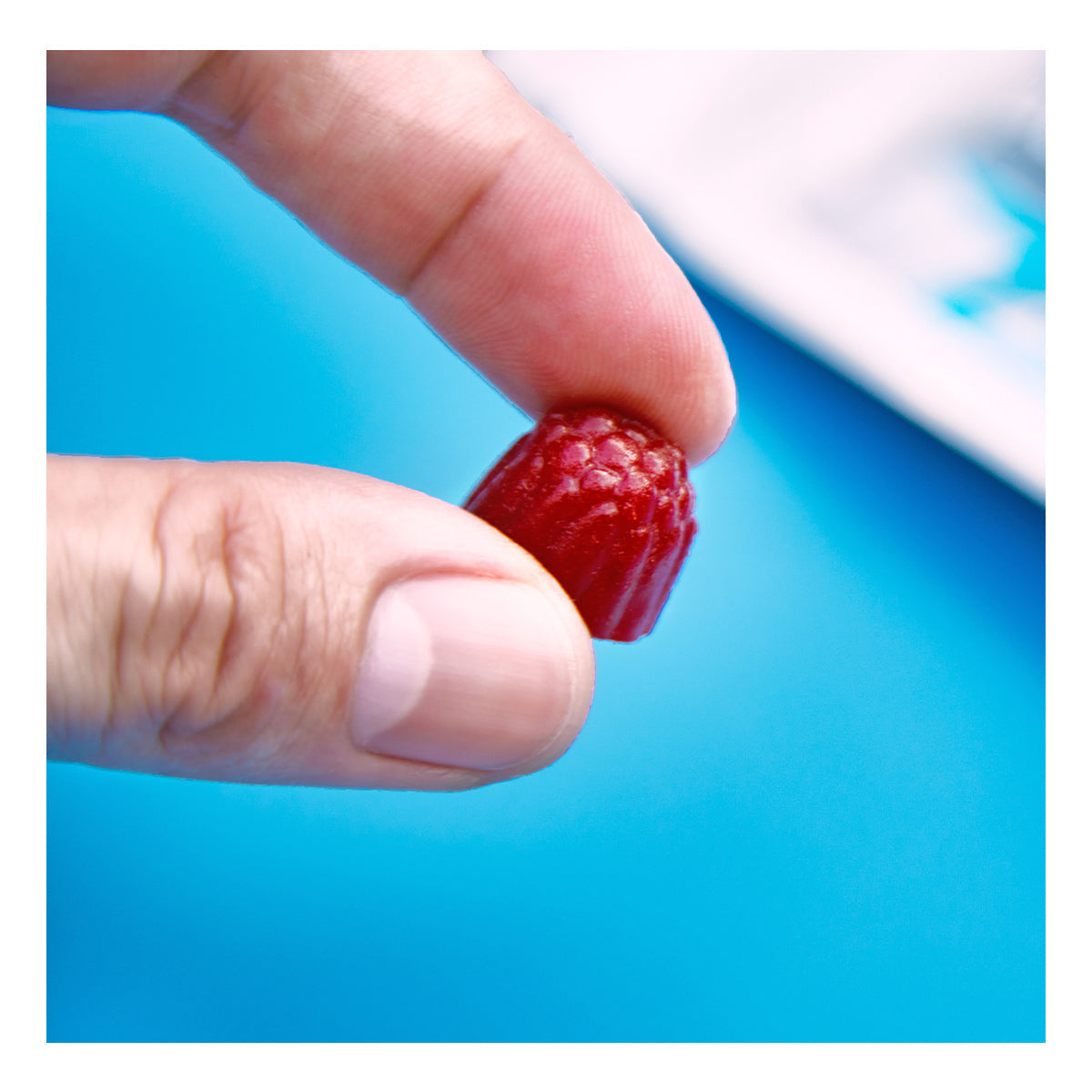 A hand holds a small, red raspberry-shaped candy against a blue background, reminiscent of FitEyez Eye Fitness Gummies (Wild Berry), which are designed with eye health nutrients.