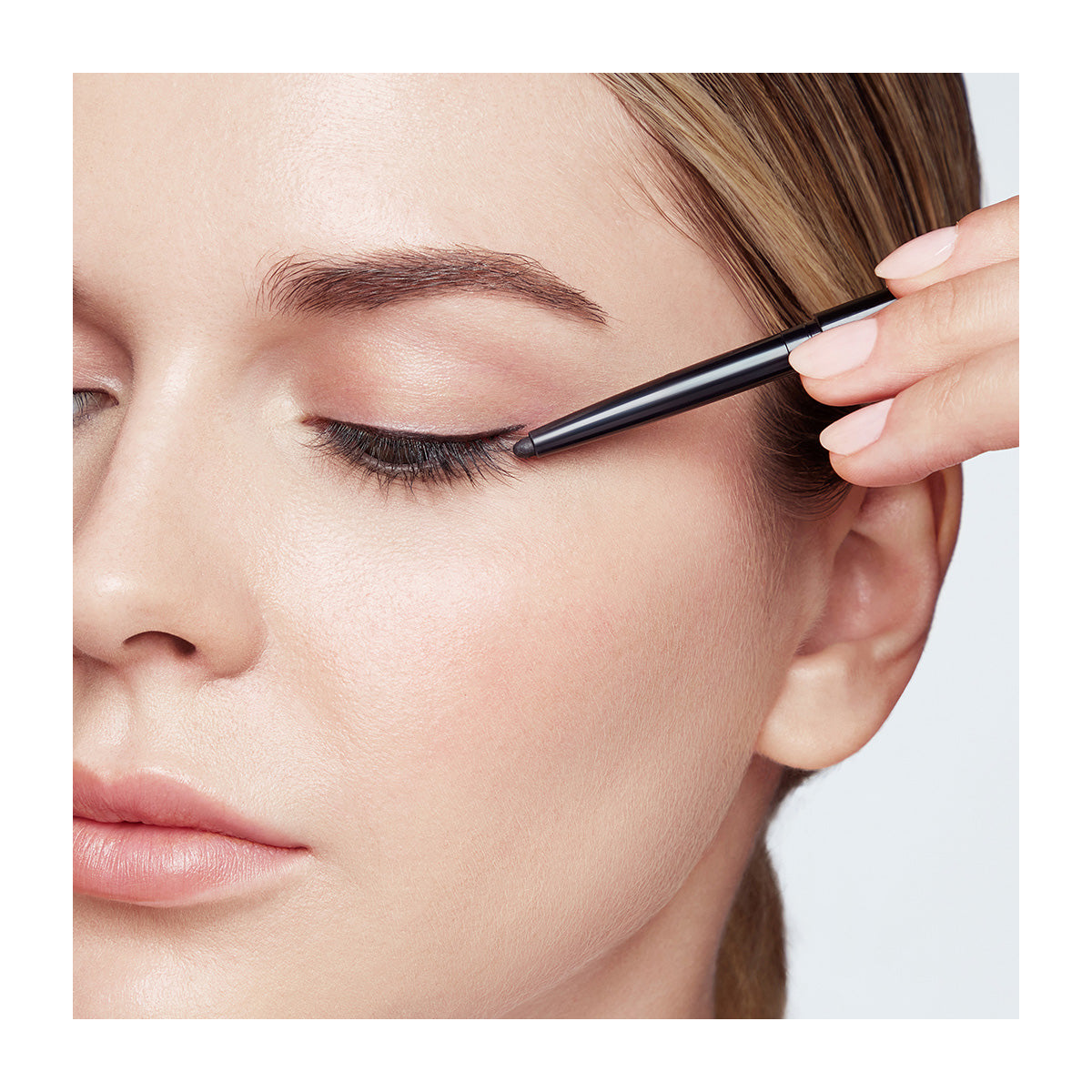 A person expertly applying RevitaLash Defining Liner Eyeliner—a smudge-proof black pencil—precisely to their upper lash line, with a focus on perfect eye makeup technique against a light background.