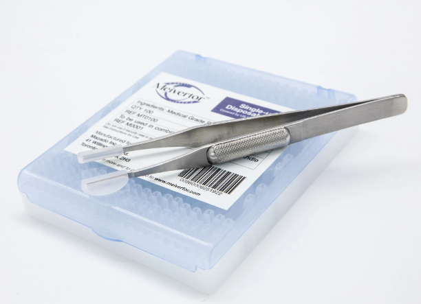 A pair of surgical forceps lies on a blue box containing the Meivertor™ Starter Kit — Stainless Steel Handle + 100 Single-Use Disposable Tips for Eyelid Eversion, suitable for procedures like Meibomian gland imaging. The box shows product details and a barcode against a plain white background.