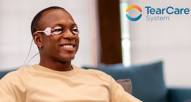 A man sits comfortably on a couch, using TearCare SmartLids Replacements by Sight Sciences to ease dry eye disease. Smiling in his beige shirt, he exudes relaxation with the Sight Sciences logo prominently displayed in the top right corner.